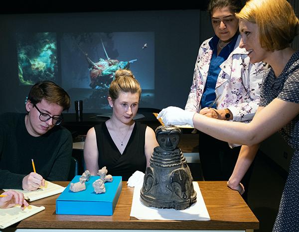Professor and students studying Pre-Columbian terracotta whistles and a 19th century wooden helmet mask from West Africa. 