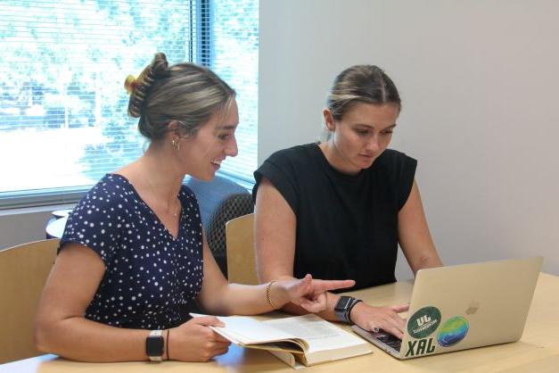 两名女研究生一边看着笔记本电脑屏幕一边交谈并互相打手势..